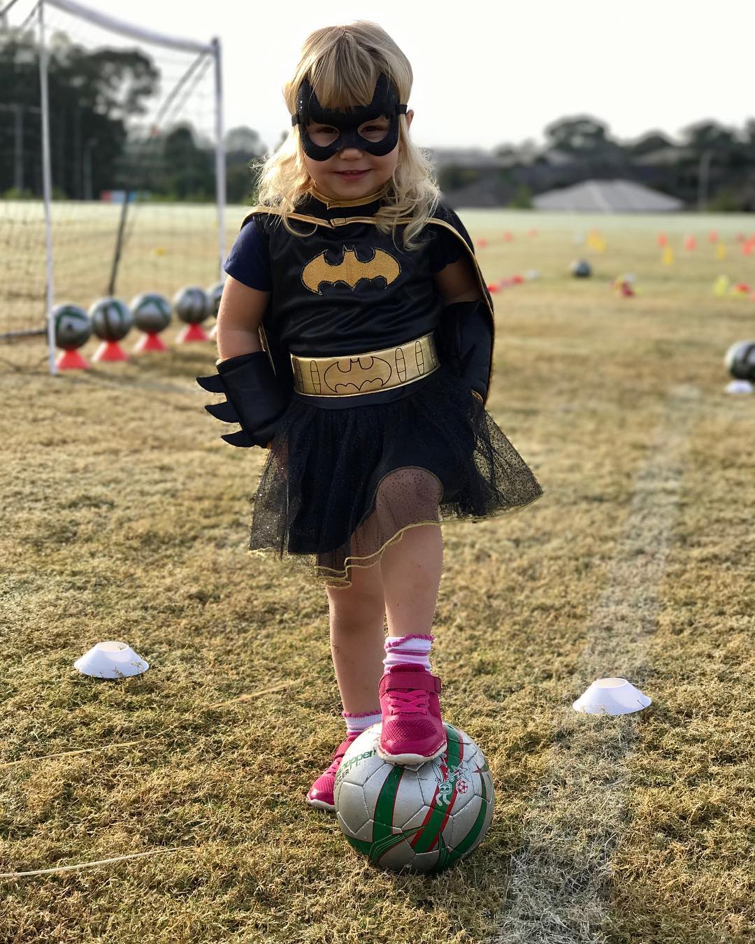 Young child in a black Batman costume and pink sneakers stands on a soccer ball on a grassy field with cones and a goal behind her.