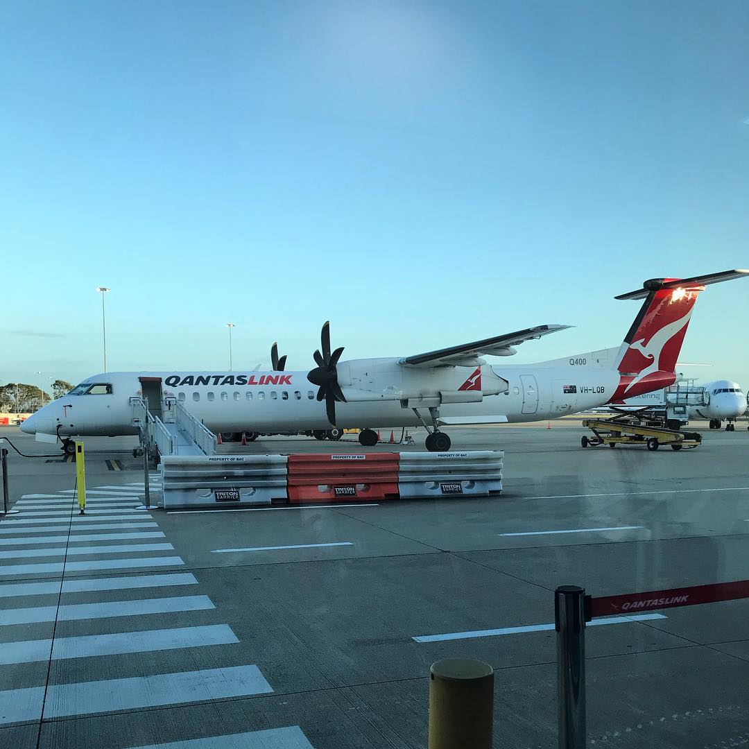 QantasLink turboprop airplane parked on the airport tarmac beside boarding stairs and ground equipment under a clear blue sky.