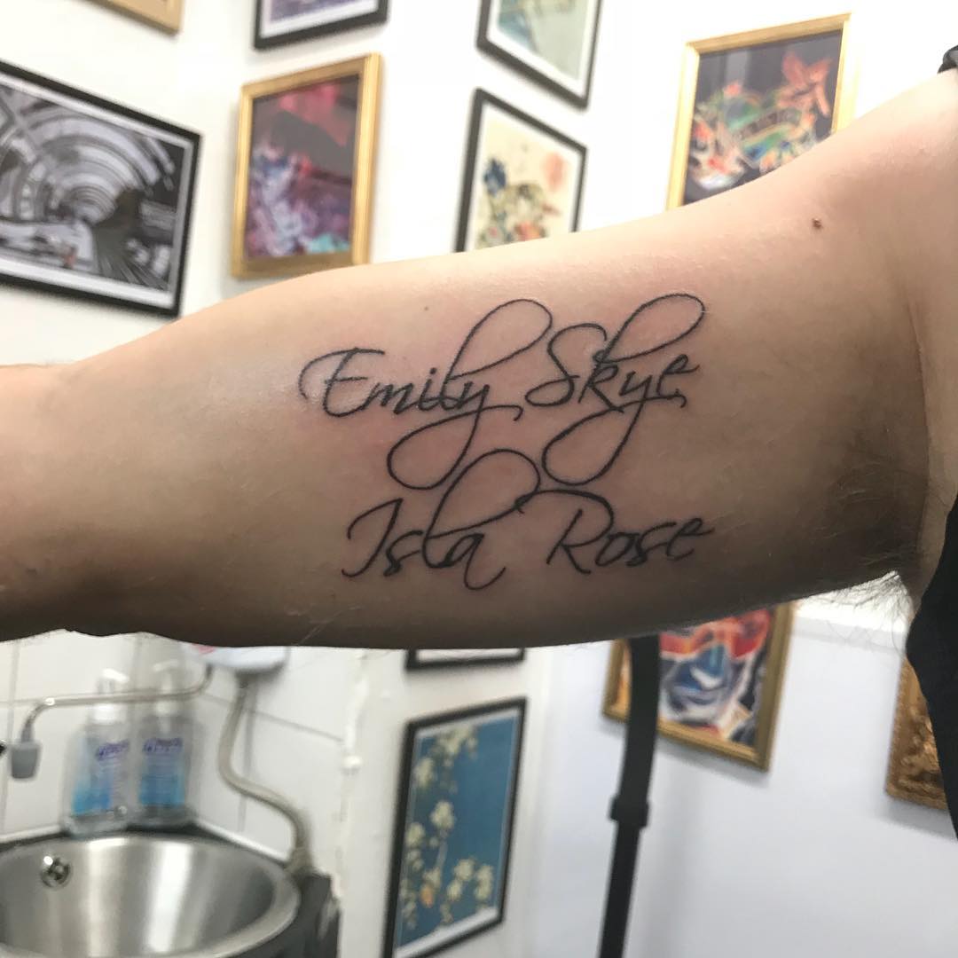 Close-up of an inner upper arm with a cursive tattoo reading “Emily Skye” above “Isla Rose,” with framed artwork and a sink in the background.