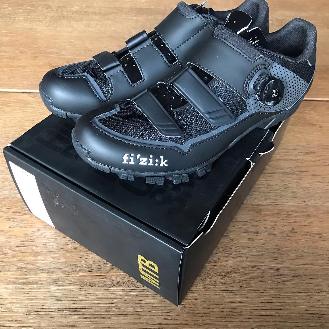 Pair of black and gray cycling shoes with Velcro straps and a side dial, resting on a black shoebox on a wooden surface.