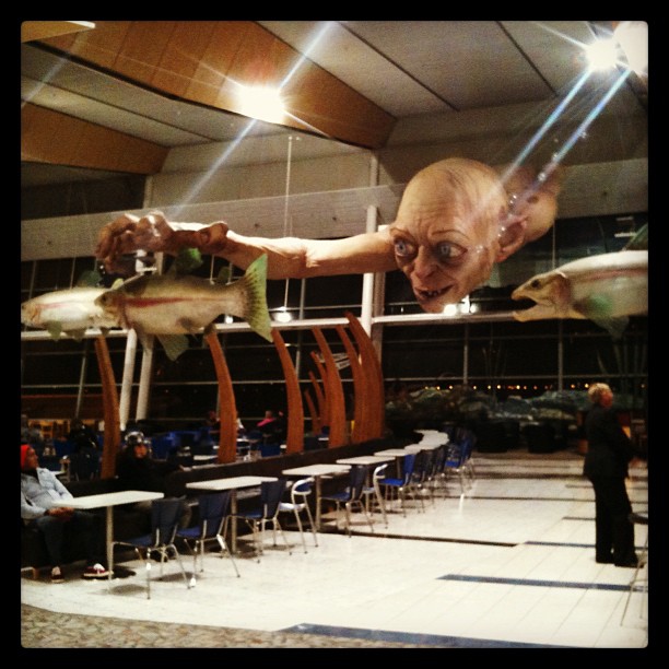 Gollum sculpture with long outstretched arms hangs above an airport seating area, holding a fish near another fish sculpture.