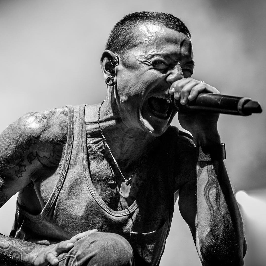 Black-and-white close-up of a tattooed singer in a tank top gripping a microphone and shouting intensely.