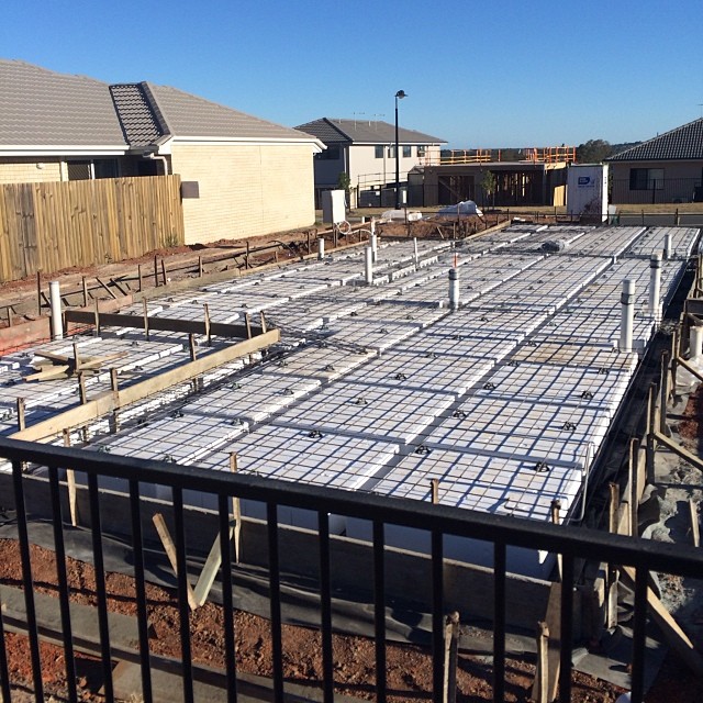 Concrete slab foundation under construction with steel mesh, white insulation panels, and plumbing pipes, bordered by nearby houses and fencing.
