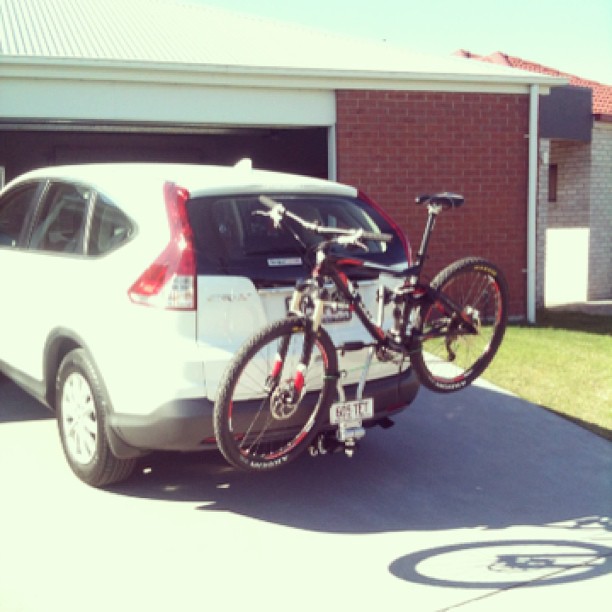 White SUV parked in a driveway with a bicycle mounted on a rear rack in front of a brick house.