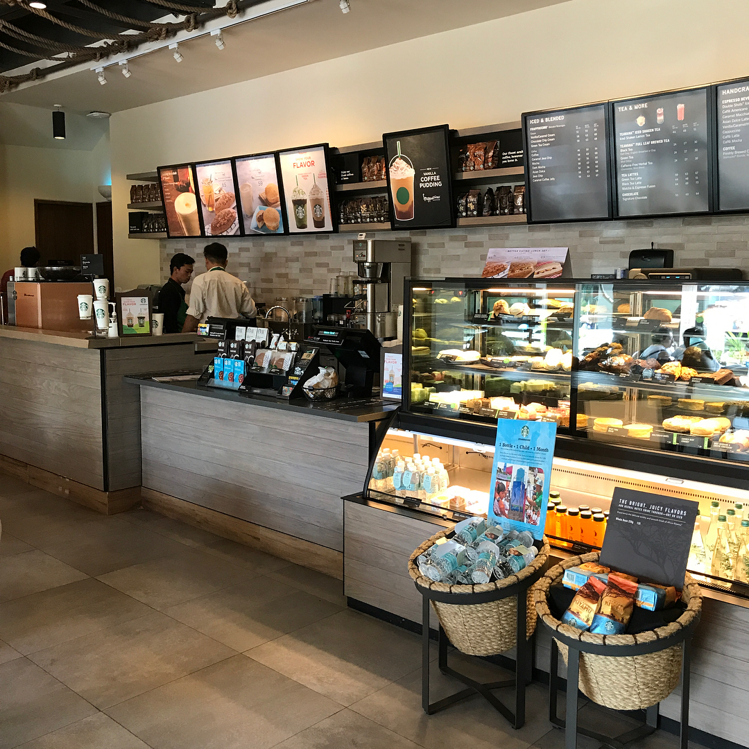 Coffee shop counter with menu boards overhead, two staff behind the register, and a glass case filled with pastries and bottled drinks.