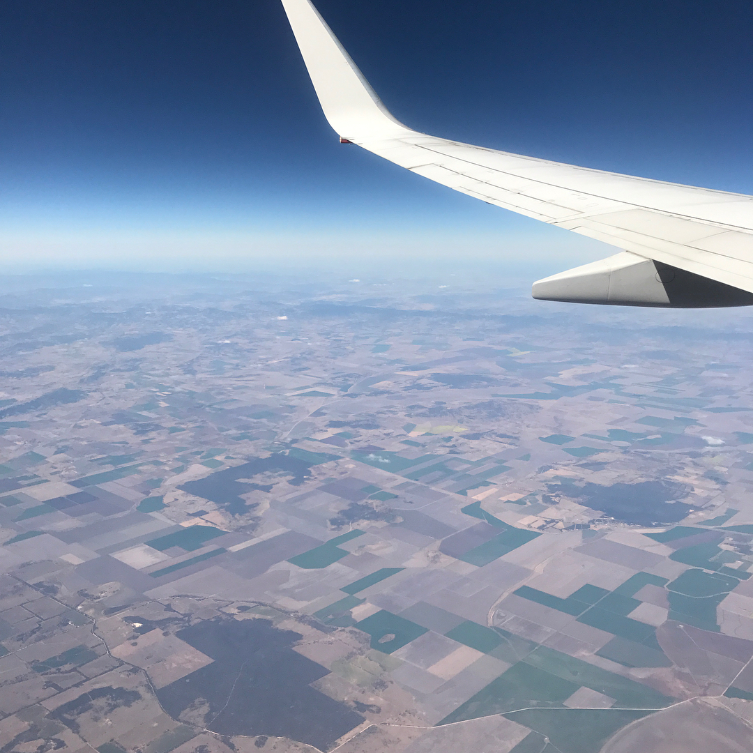 Airplane wing above a patchwork of fields seen from high altitude under a clear blue sky.