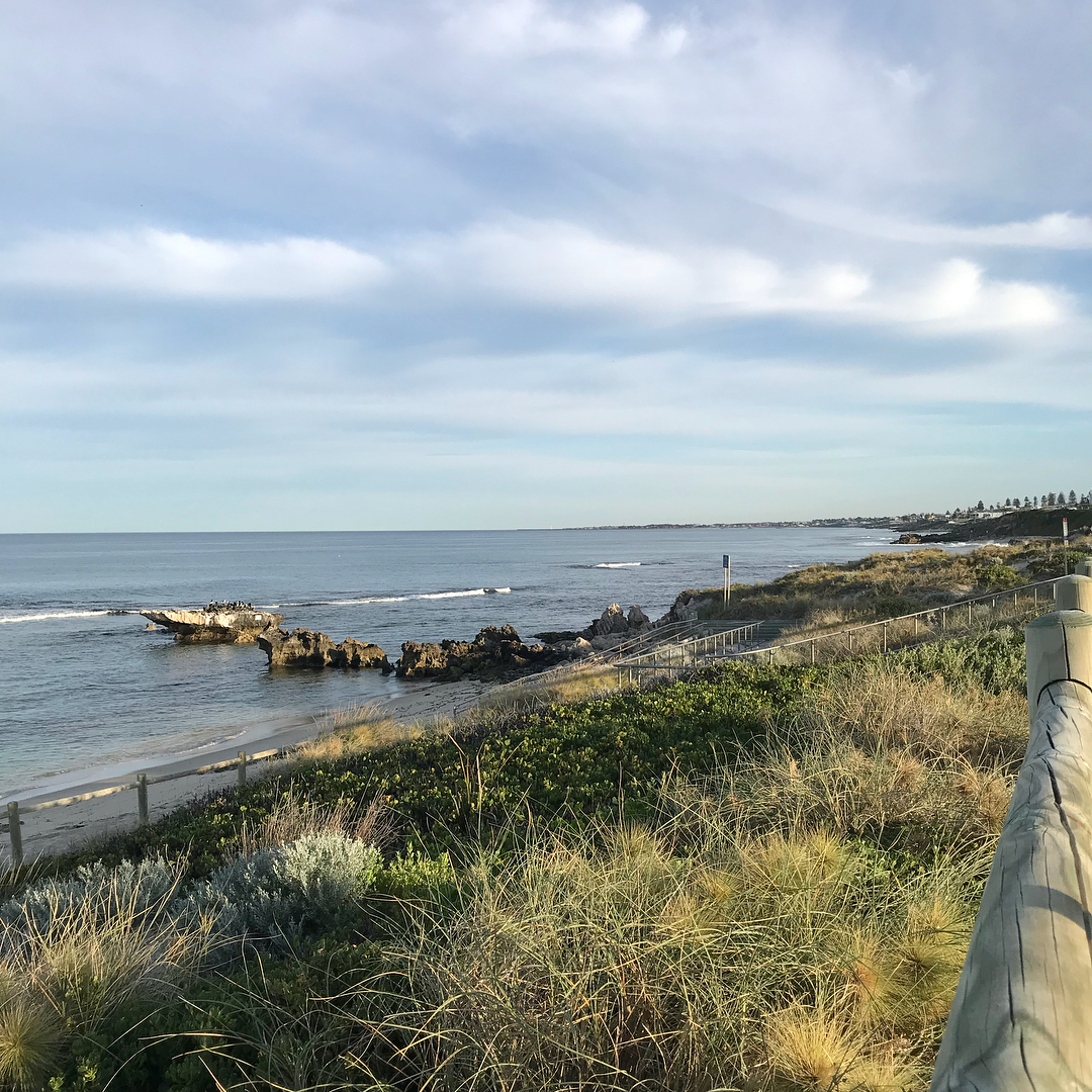 Calm coastal shoreline with rocky outcrops, gentle waves, grassy dunes, and a path railing beneath a wide cloudy sky.