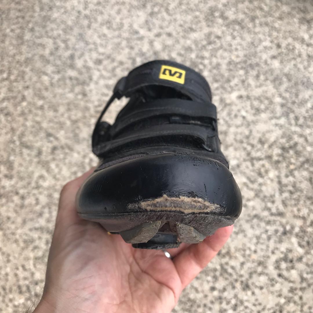 Hand holding a black cycling shoe with worn, damaged toe and sole against a rough concrete background.