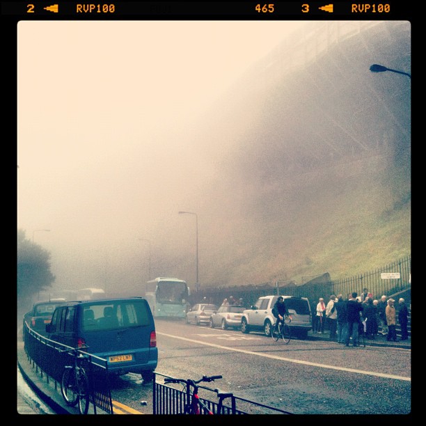 Foggy street with parked cars, a bus, cyclists, and a crowd on the sidewalk beside a steep hillside.