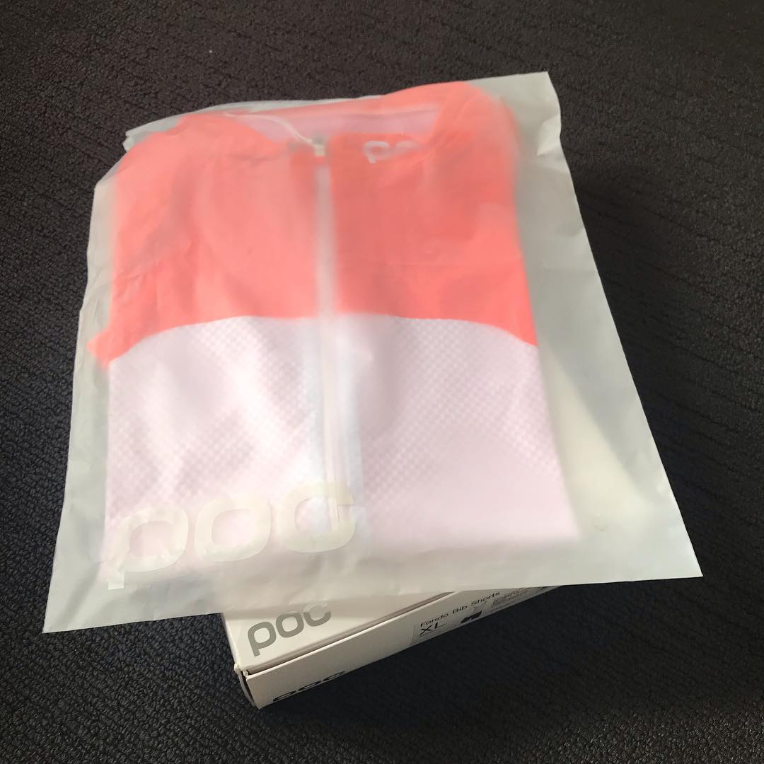 Bright pink and white zip-front jersey inside a translucent bag, resting on a white box on dark fabric.