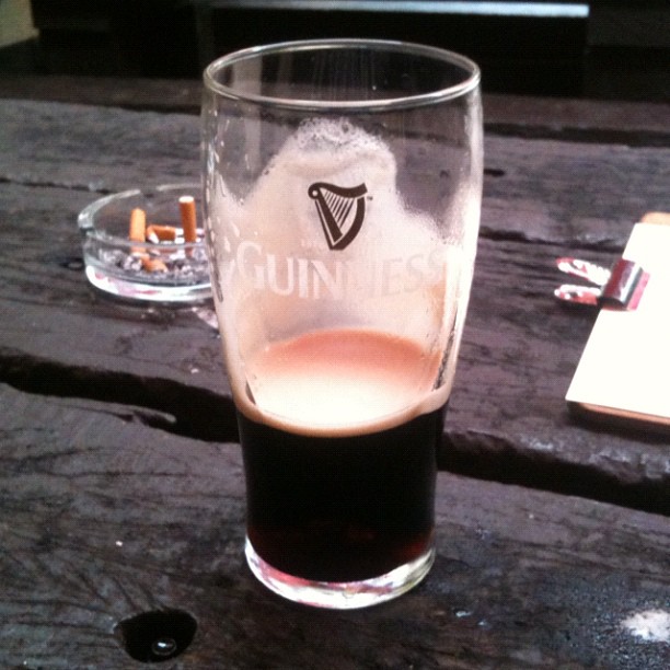 Half-full Guinness glass on a dark wooden table, with an ashtray and cigarette butts in the background.