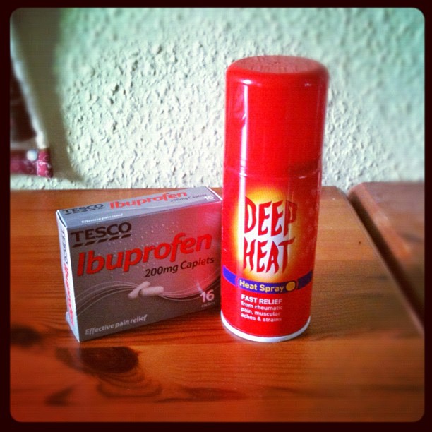 A box of Tesco ibuprofen caplets beside a can of Deep Heat spray on a wooden table.