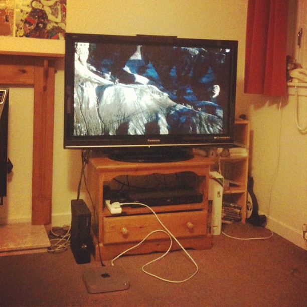 Television on a wooden stand in a living room, with cables on the carpet and shelves holding a game console and discs.