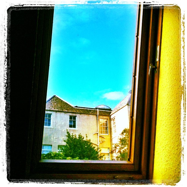 Open window framing pale buildings, green trees, and a bright blue sky.