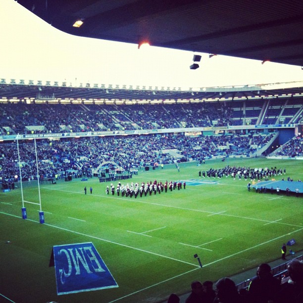 Crowded rugby stadium with goalposts and marching bands lined up on the field before a ceremony.