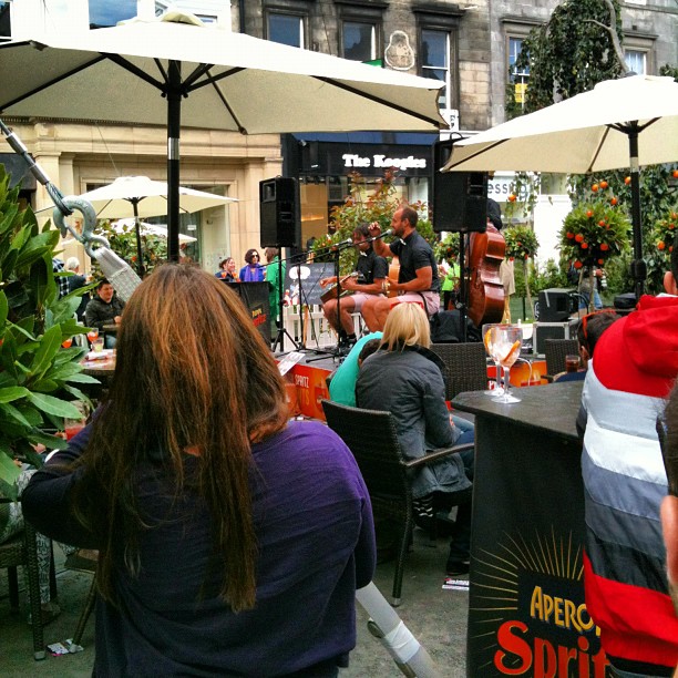 Outdoor café audience watching musicians perform under large white umbrellas in a city square.