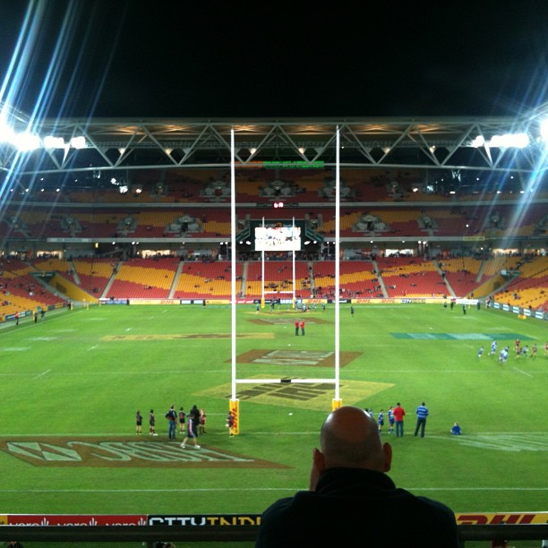 Rugby field under stadium lights with goalposts centered, red and yellow seats, scattered people on the grass, and a spectator in the foreground.