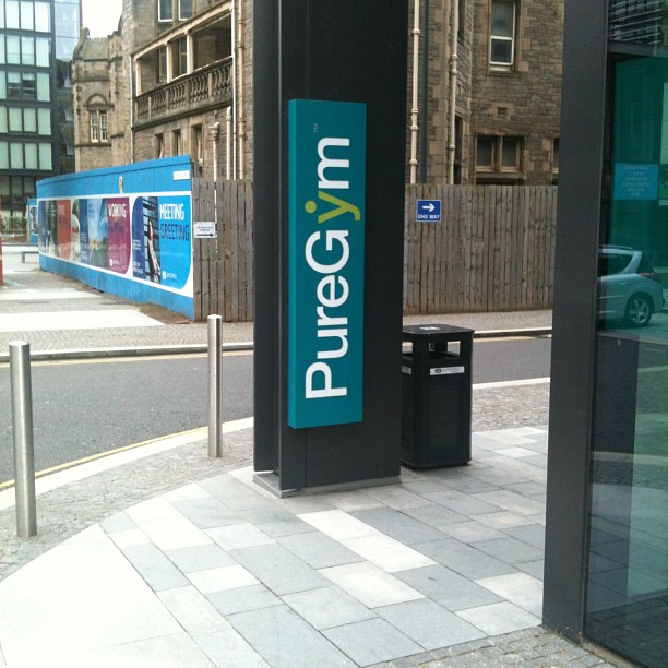 Vertical PureGym sign outside a building entrance on a city street, with bollards, a bin, and stone buildings in the background.