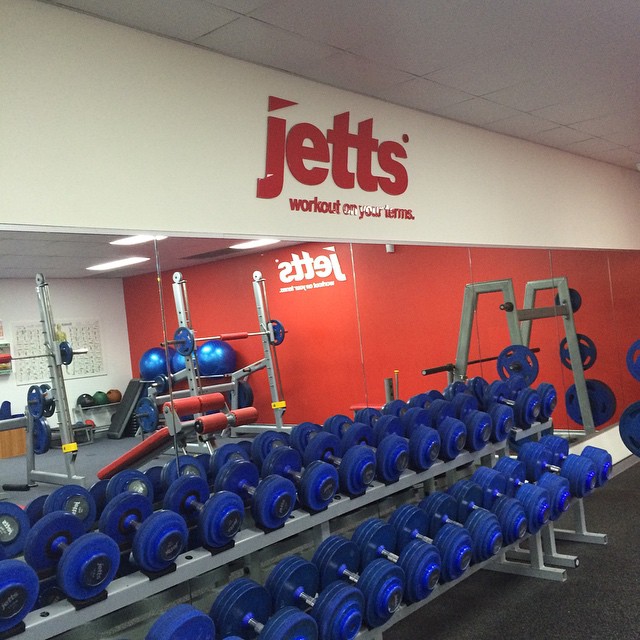Rows of blue dumbbells and gym equipment in front of a mirrored wall with a red “jetts” sign above.