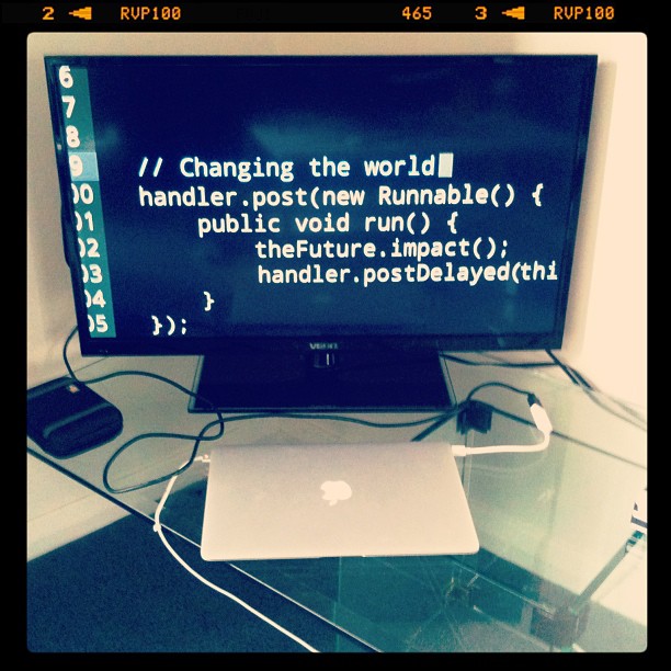 Monitor displaying code above a closed laptop on a glass desk, with cables and small devices on either side.