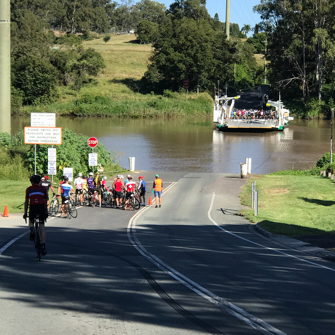 Cyclists wait on a riverside road for a ferry carrying more cyclists across the water.
