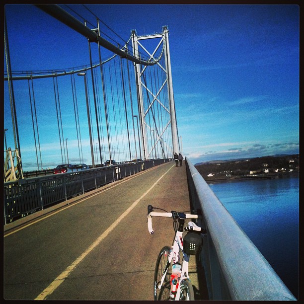 White road bike parked on the sidewalk of a suspension bridge above blue water, with cars on the road and two people ahead.
