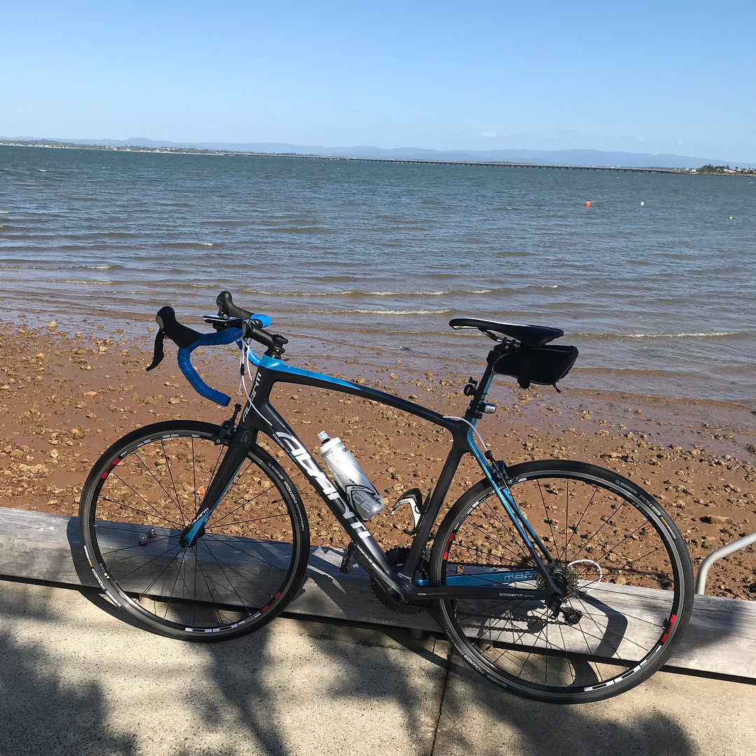 Black and blue road bike parked by a rocky sandy shore with calm water and distant hills under a clear blue sky.
