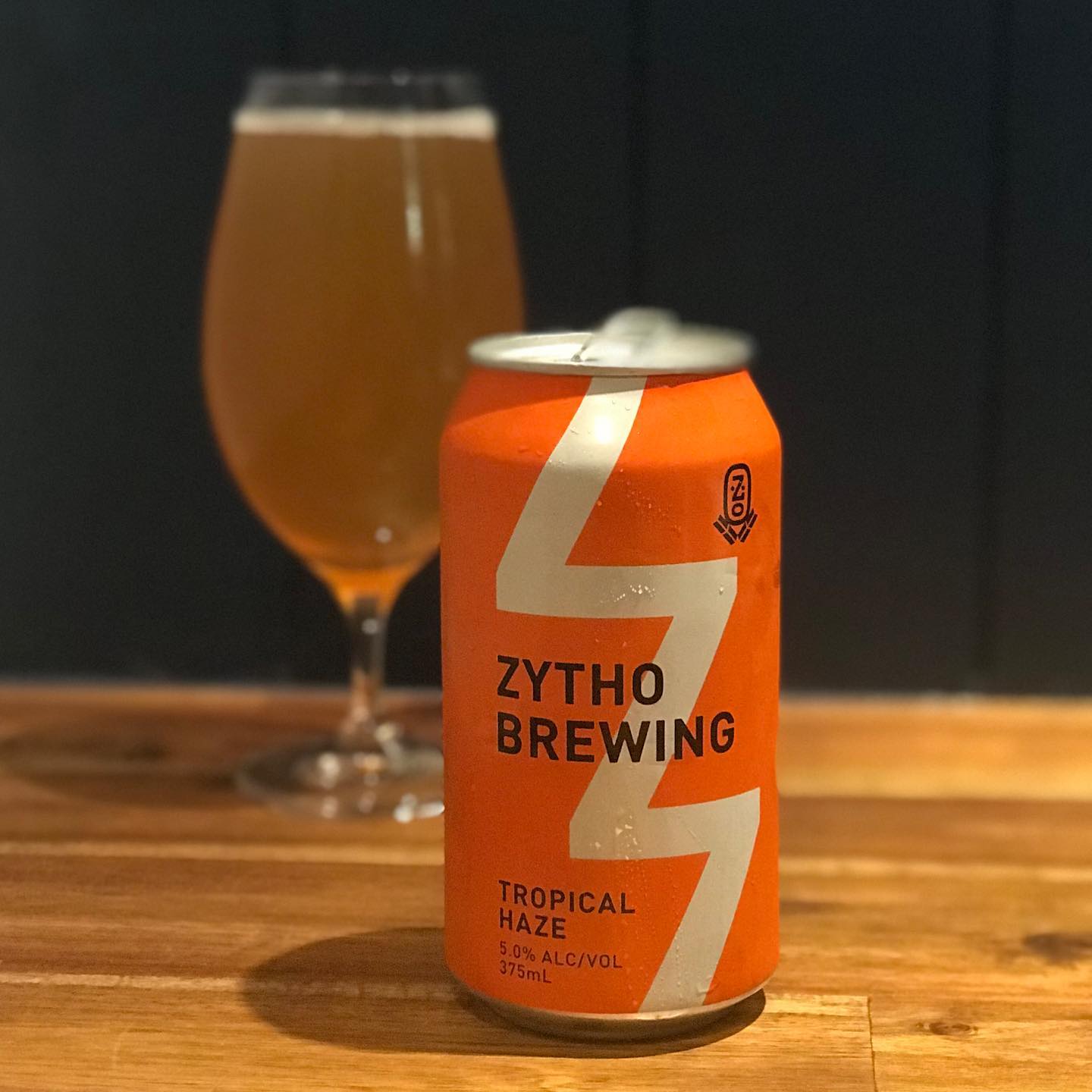 Orange Zytho Brewing Tropical Haze beer can on a wooden table, with a filled tulip glass blurred in the background.