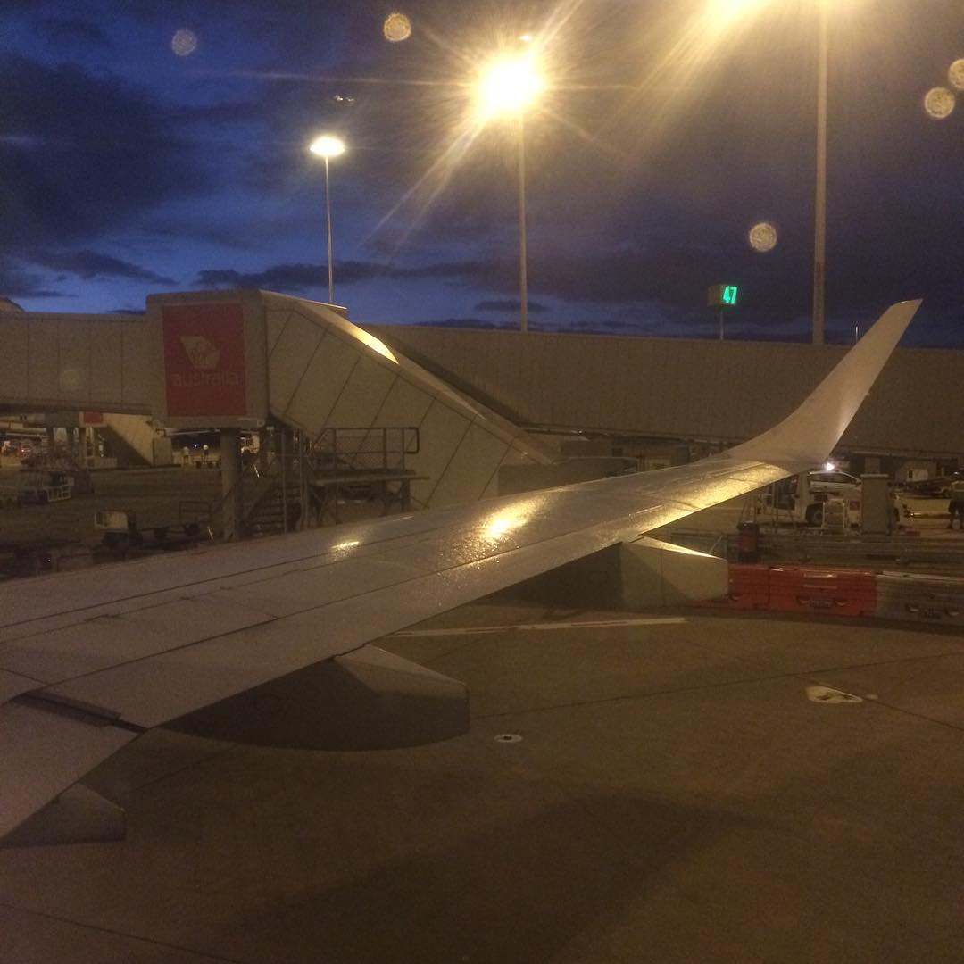 View from an airplane window of a wing at the gate at night, with jet bridge, bright lights, and a dark blue sky.