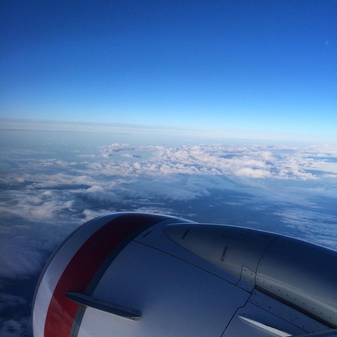 View from an airplane window showing an engine in the foreground above a blanket of clouds under a clear blue sky.