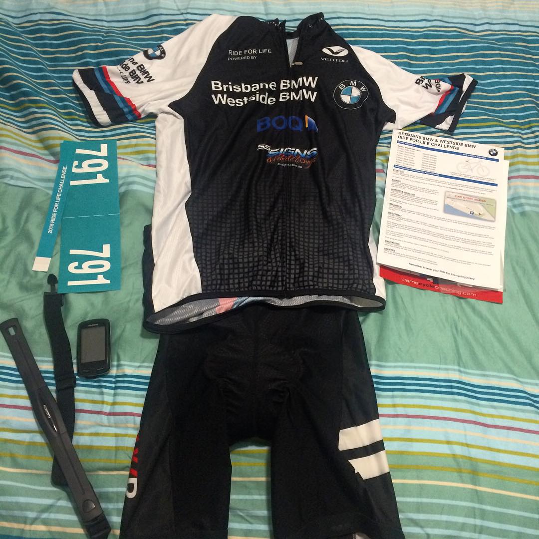 Cycling jersey and shorts laid out on a striped bedspread with race bib 791, a bike computer, heart rate strap, and event papers.