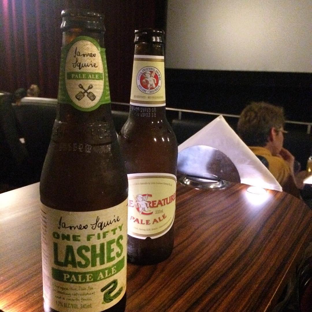 Two bottles of pale ale on a wooden table in a dim theater, with seats, a folded napkin, and people in the background.
