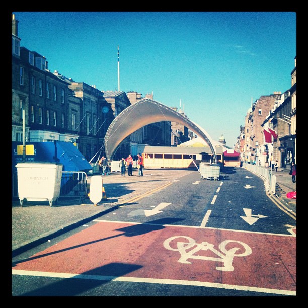 City street with a red bike lane, barriers, and a large arched temporary structure between rows of buildings under a clear blue sky.