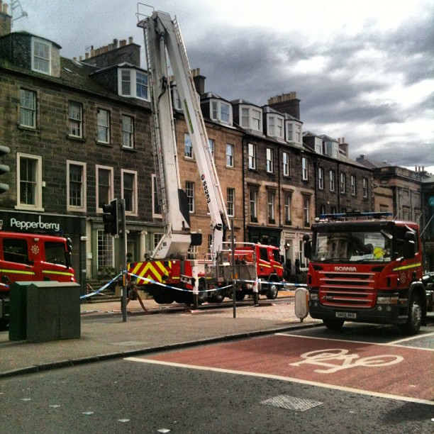 Fire trucks and a raised ladder parked on a city street beside stone row buildings under a cloudy sky.
