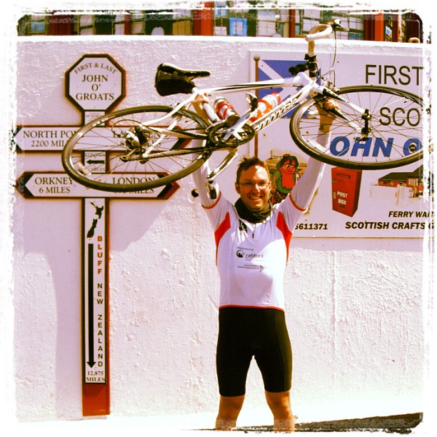 Cyclist in riding gear lifts a road bike overhead in front of a white wall with John O’Groats signs and a large route marker.