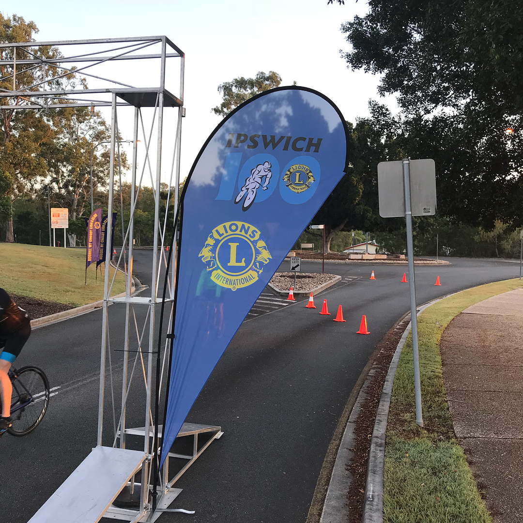 Blue Ipswich Lions cycling event banner beside a road with orange cones, trees, and a cyclist passing on the left.