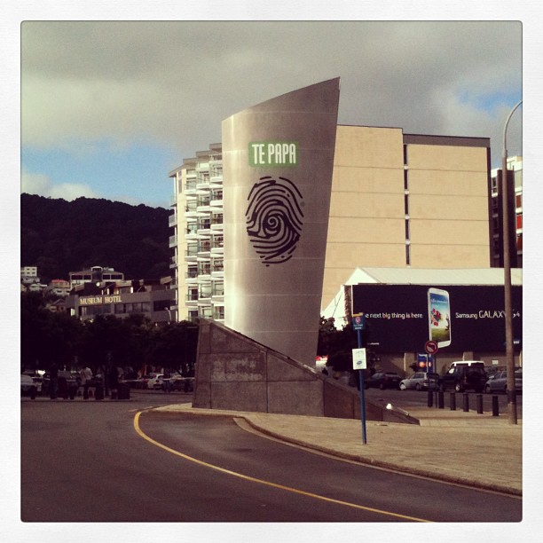Curved silver Te Papa sign with a fingerprint logo beside a road, with buildings, cars, and cloudy sky in the background.