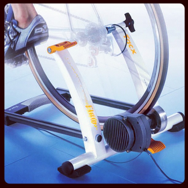Bicycle rear wheel mounted on an indoor trainer, with a cyclist’s shoe on the pedal and orange adjustment parts visible.