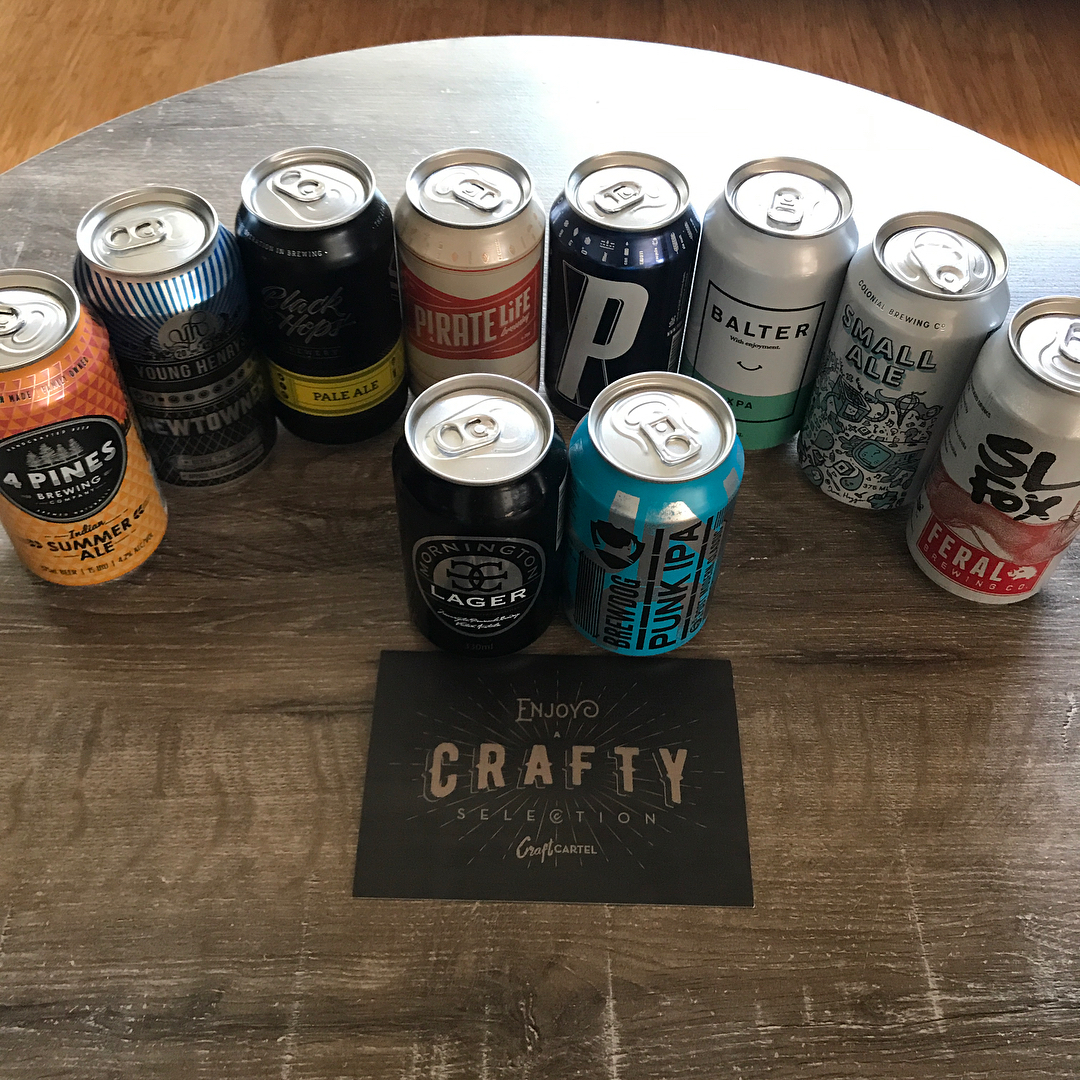 Nine assorted beer cans arranged on a wooden table around a card reading “Enjoy a Crafty Selection.”