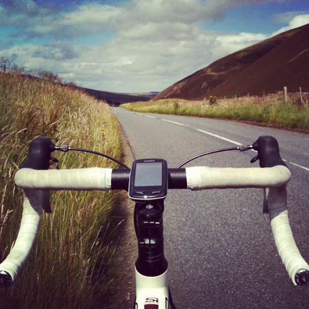 View over white-wrapped bicycle handlebars and bike computer on a paved road between grassy hills under a cloudy sky.