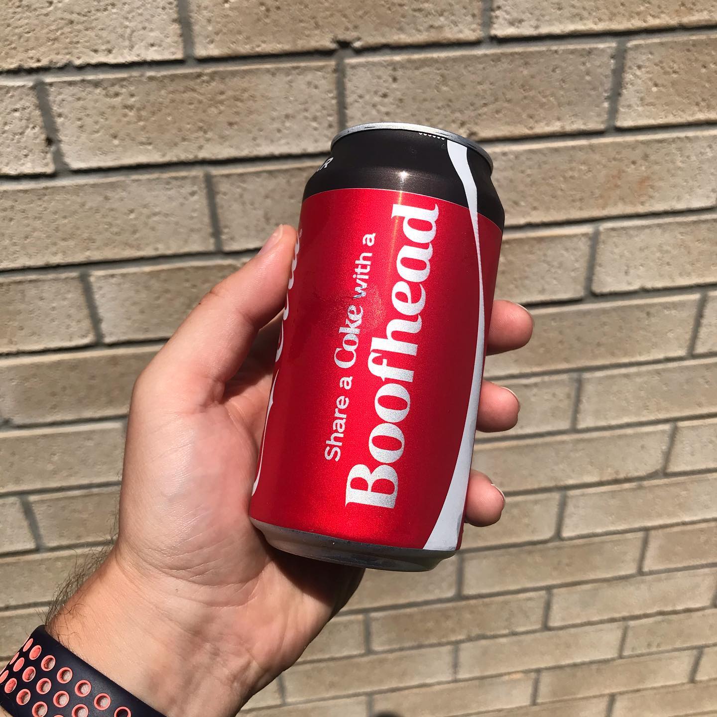 Hand holding a red Coca-Cola can labeled “Share a Coke with a Boothead” in front of a tan brick wall.