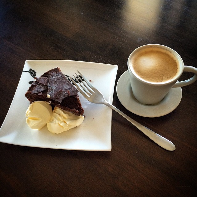 Slice of chocolate cake with cream on a white plate and a fork beside a cup of coffee on a dark wooden table.