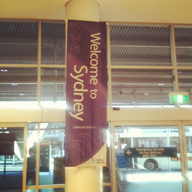 Purple banner on a pillar reading “Welcome to Sydney” inside an airport entrance with glass doors and a lit exit sign.