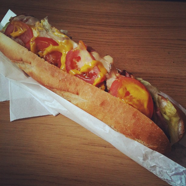 Hot dog in a long bun with tomato slices and mustard, wrapped in white paper on a wooden table.