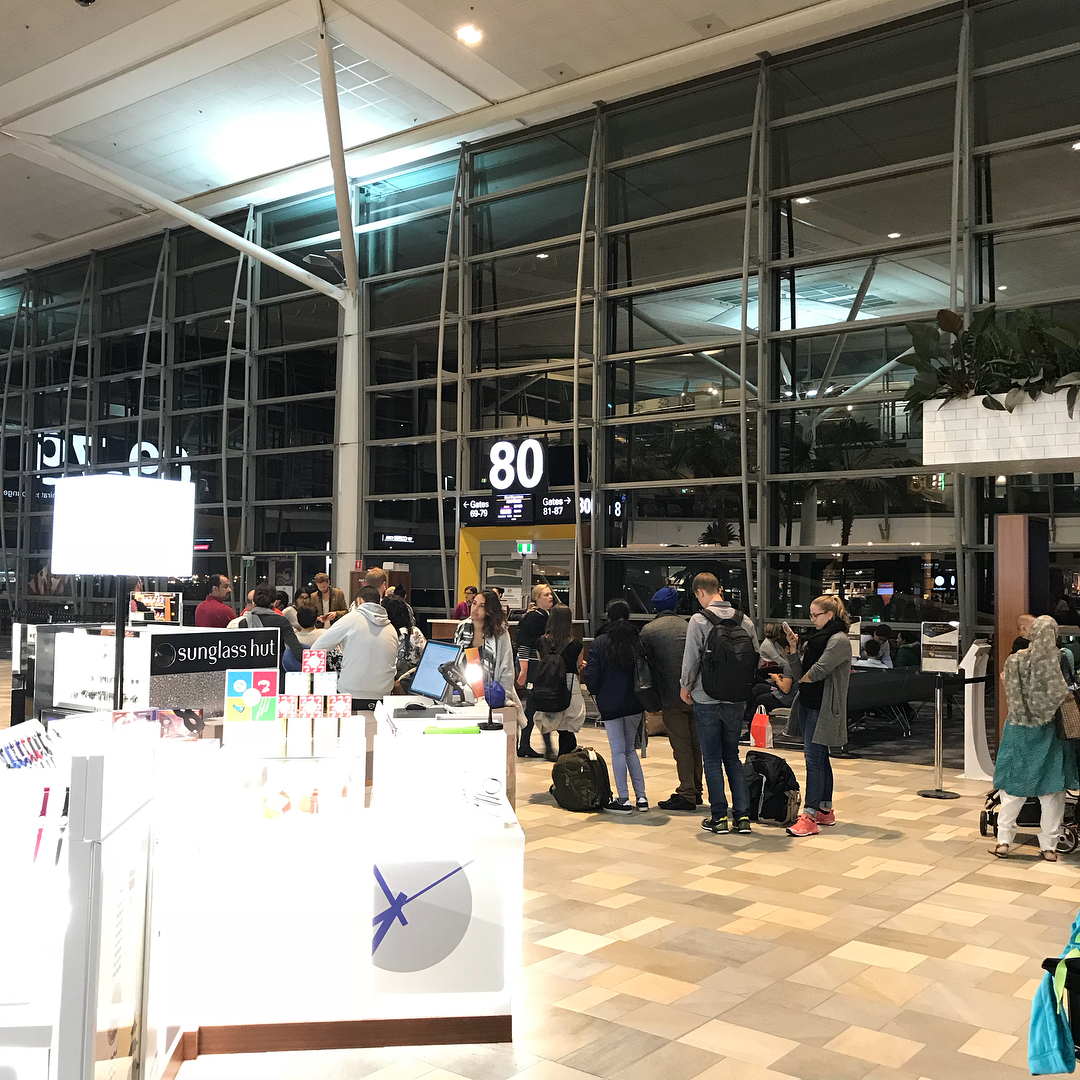 Passengers wait near gate 80 in a brightly lit airport terminal beside a Sunglass Hut kiosk and large glass windows.