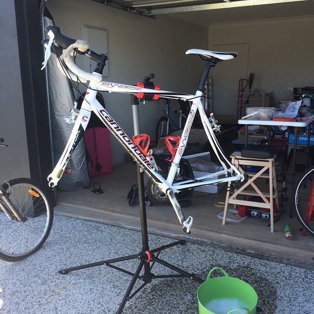 White Cannondale road bike frame mounted on a repair stand in a garage, with wheels removed and tools and items in the background.