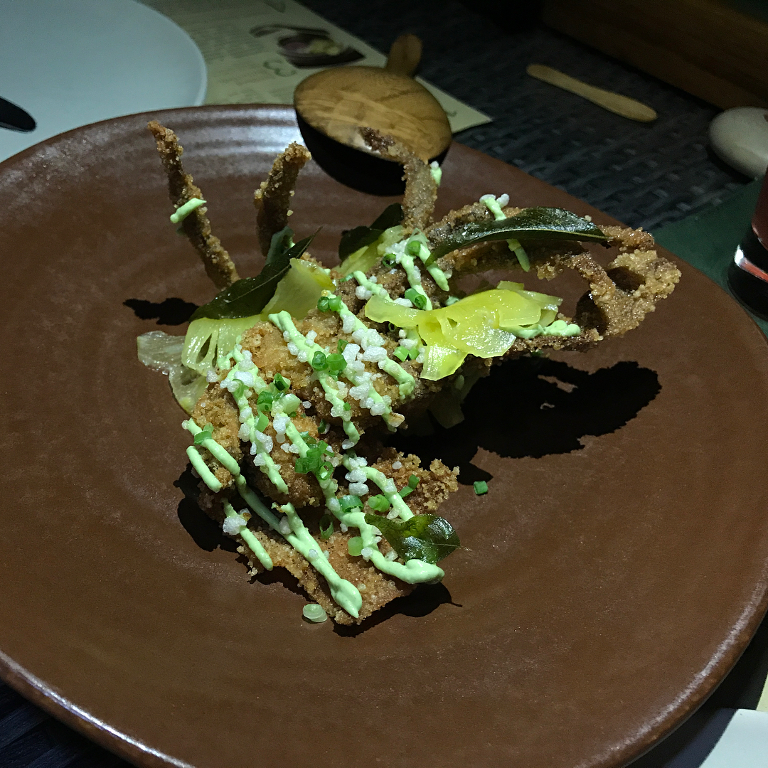Breaded fried pieces on a brown plate, topped with green sauce, chopped scallions, coarse salt, and pale yellow vegetable slices.