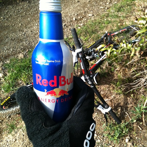 Gloved hand holding a blue Red Bull bottle in front of a bicycle lying on a dirt path.