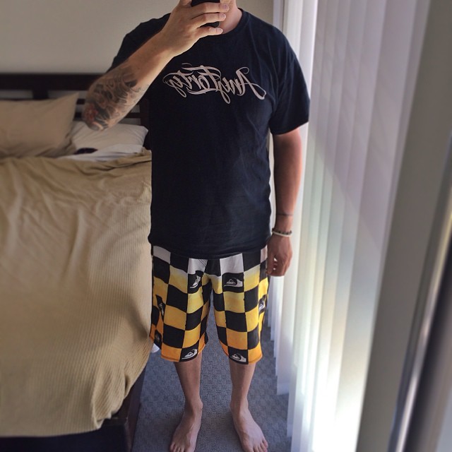 Person taking a mirror selfie in a bedroom, wearing a black T-shirt and yellow-and-black checkered shorts, barefoot beside a bed and window blinds.