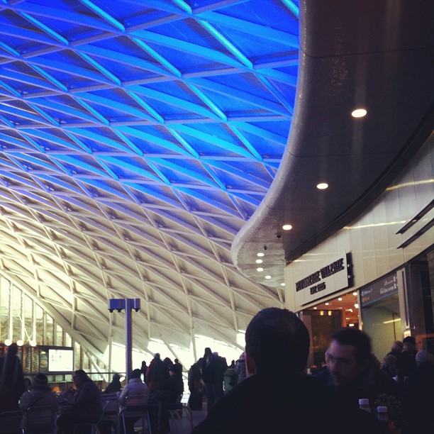 People sit and walk beneath a large curved ceiling with blue geometric lighting inside a spacious terminal.